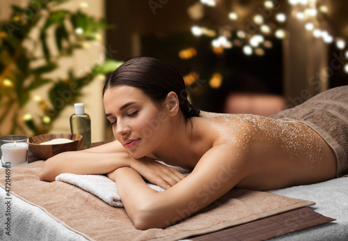 wellness, beauty and relaxation concept - beautiful young woman lying with sea salt on skin at spa over garland lights on window background