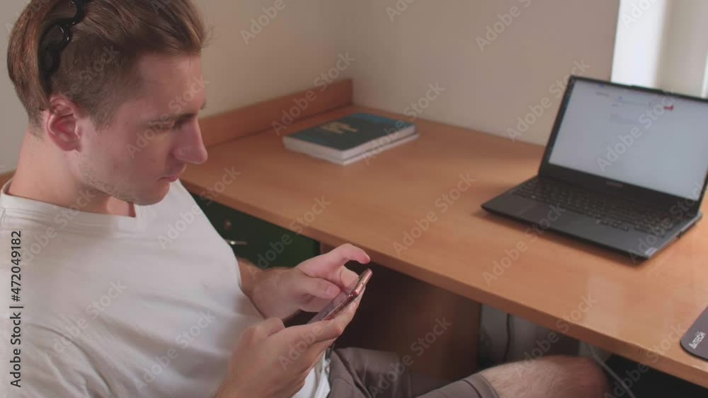 Student guy uses mobile internet in the phone at the table. He points his fingers at the screen. There is a laptop in bokeh on the table. Student guy uses mobile internet in the phone at the table. He points his fingers at the screen. There is a laptop in bokeh on the table.
