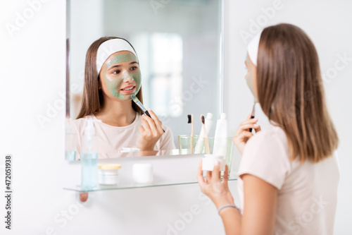 beauty  skin care and people concept - teenage girl with brush applying clay mask to face and looking in mirror at bathroom