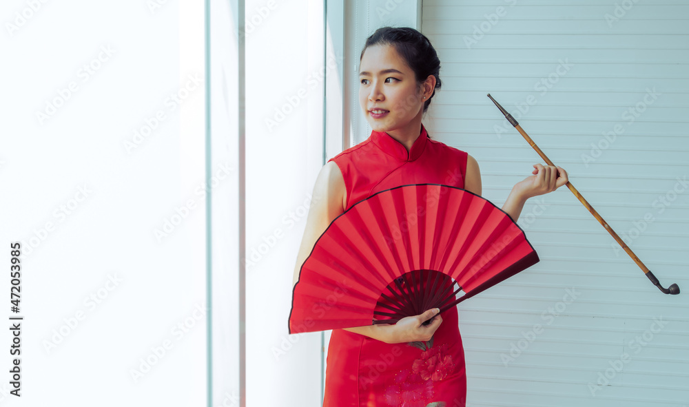 Young Asian Chinese beautiful woman wearing red traditional dress, holding folding fan and pipe, standing beside window in a day at indoor room, smiling to celebrate new year with copy space. Young Asian Chinese beautiful woman wearing red traditional dress, holding folding fan and pipe, standing beside window in a day at indoor room, smiling to celebrate new year with copy space.