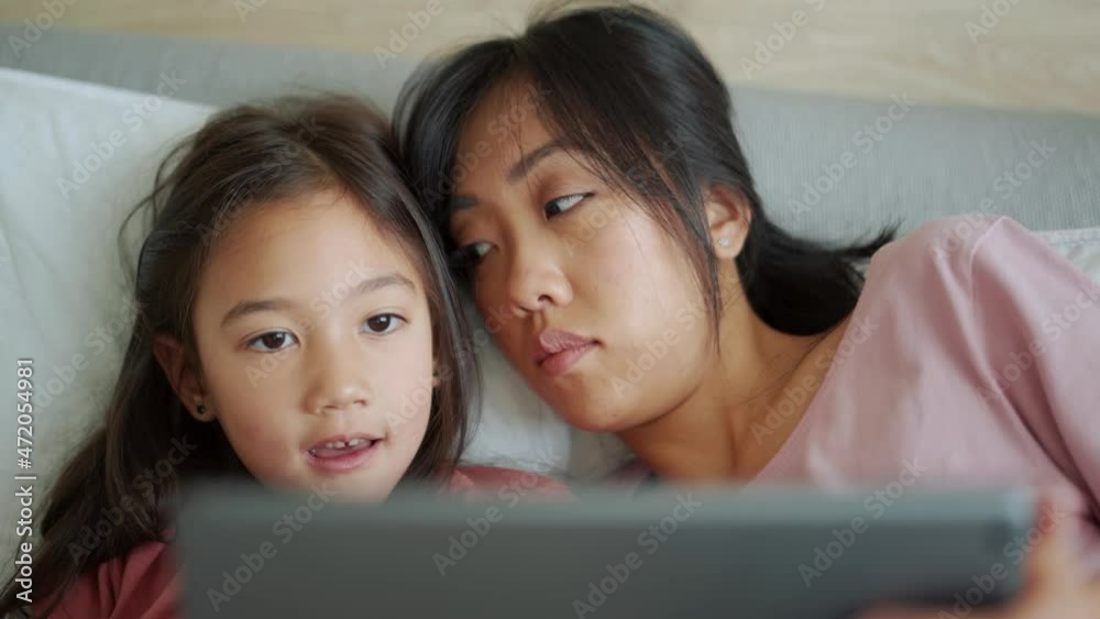 Pensive Asian mother and daughter looking at the tablet and talking on the bed Pensive Asian mother and daughter looking at the tablet and talking on the bed
