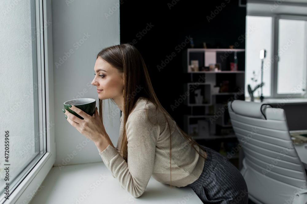 Businesswoman with coffee mug leaning on window sill in office Businesswoman with coffee mug leaning on window sill in office
