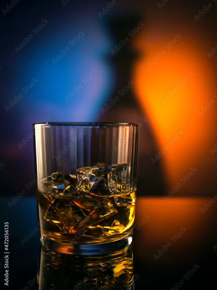 Whiskey, cognac, bourbon in a glass with ice on a mirrored table, colorful background. Whiskey, cognac, bourbon in a glass with ice on a mirrored table, colorful background.