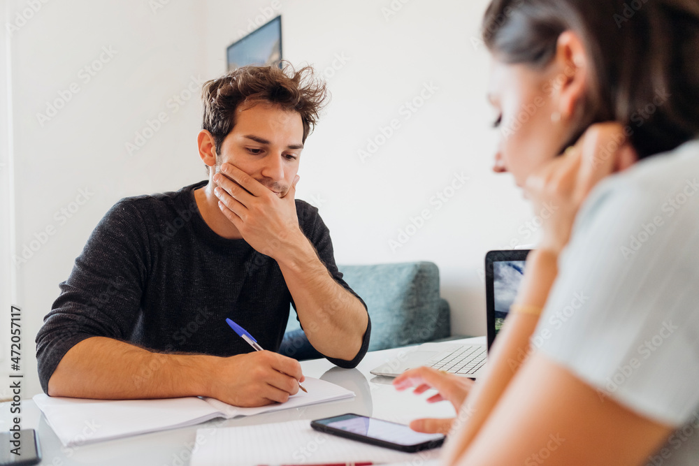 Young man with book and pen looking at businesswoman working on smart phone at home Young man with book and pen looking at businesswoman working on smart phone at home