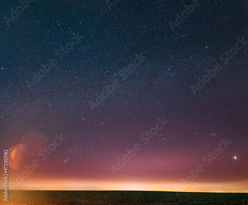 Night Starry Sky With Glowing Stars Above Countryside Landscape. Milky Way Galaxy And Rural Field Meadow In Early Spring. Bright Glow Of Planet Venus Among The Stars. Warm Lights Of Evening Sunset