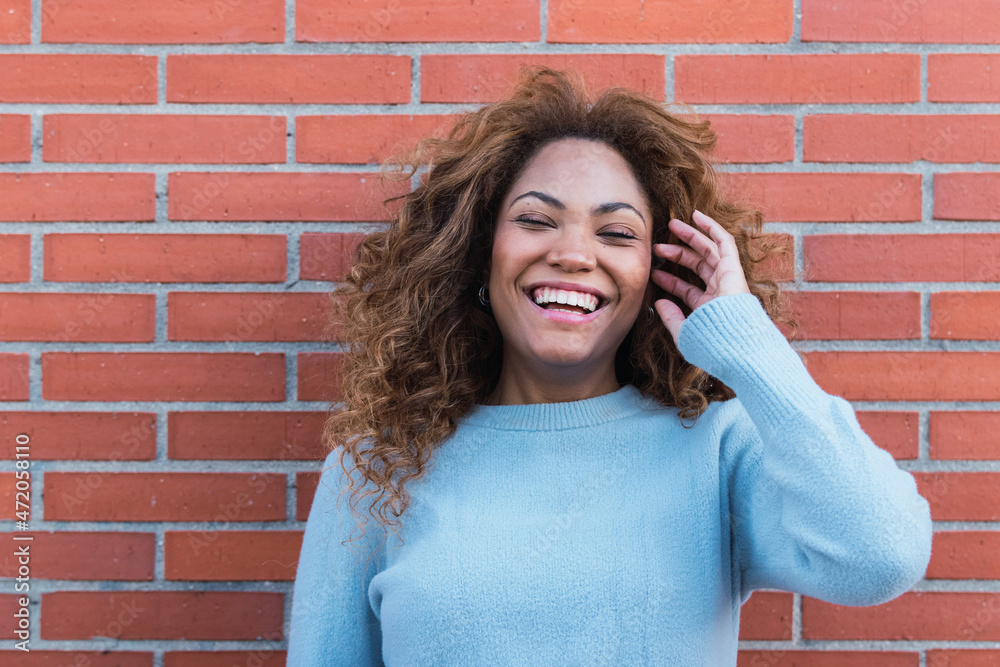 Smiling african american woman with white perfect smile, long curly hair, glamour makeup and blue jumper posing in brick wall. Beauty woman Latin Ethnic female face. Dentist concept Smiling african american woman with white perfect smile, long curly hair, glamour makeup and blue jumper posing in brick wall. Beauty woman Latin Ethnic female face. Dentist concept