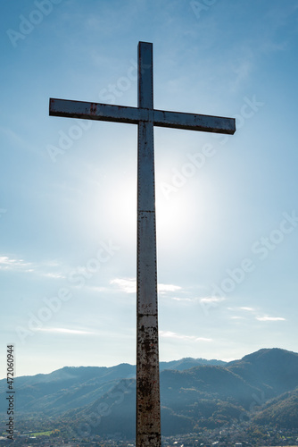 Silhouette of an iron cross at the summit of a hill where stands the "Castello dell'Innominato" (meaning: castle of the man without name), Lecco, Italy. Moutain range and blue sky on the background.
