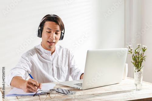 Smart Asian young businessman takes a note wearing headphones working at home office pleased happy cheerful on online meeting video or learning by a laptop computer for job work or makes good dealing