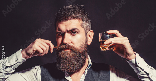 Man with beard holds glass of brandy. Tasting and degustation concept. Bearded businessman in elegant suit with glass of whiskey. Man drinking whiskey, brandy, cognac. Degustation, tasting