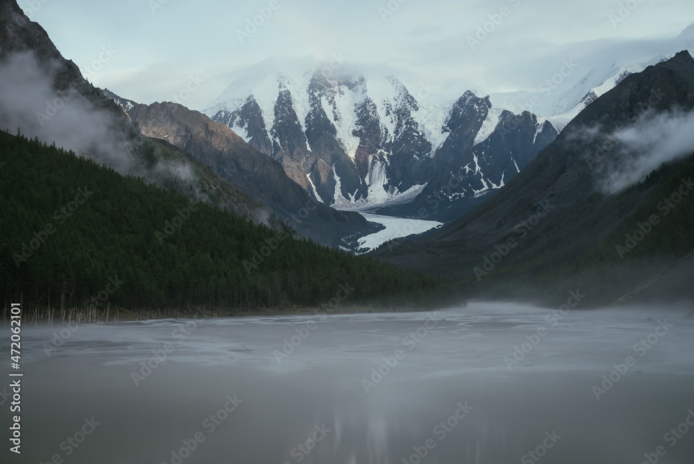 Atmospheric misty alpine landscape with gray mountain lake in fog and snowy mountains in overcast weather. Gloomy foggy scenery with mountain lake and mountain streams from glacier and low clouds.