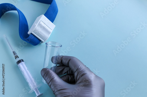 A hand in a gray medical glove holds an empty test tube, a syringe and a tourniquet for taking blood from a vein are lying next to it. Light blue background, space for text photo