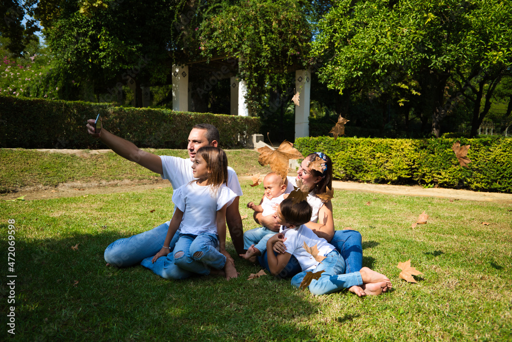 Obraz premium family portrait of five members, father, mother and children sitting on the grass taking a photo with the mobile phone while the leaves fall from the trees in autumn. Family concept.