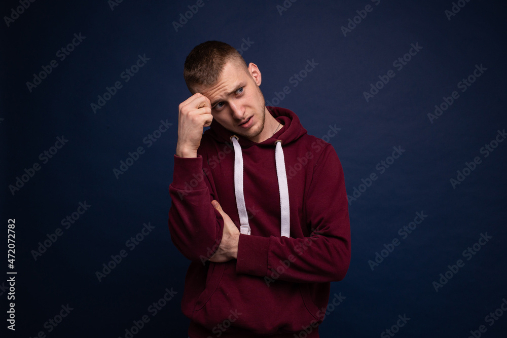 Young man in red hoodie and jeans posing on a blue background, thinking and looking away