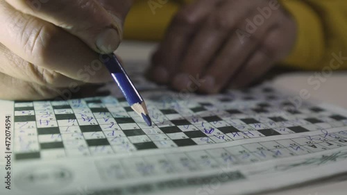 Pensioner in bright yellow sweater fills out crossword puzzle thoughtfully and writes words with blue pencil extreme closeup photo