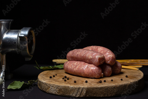 Raw meat sausages on a wooden board on a black background. Homemade sausages in a natural casing next to a steel meat grinder. Meat products. Home cooking