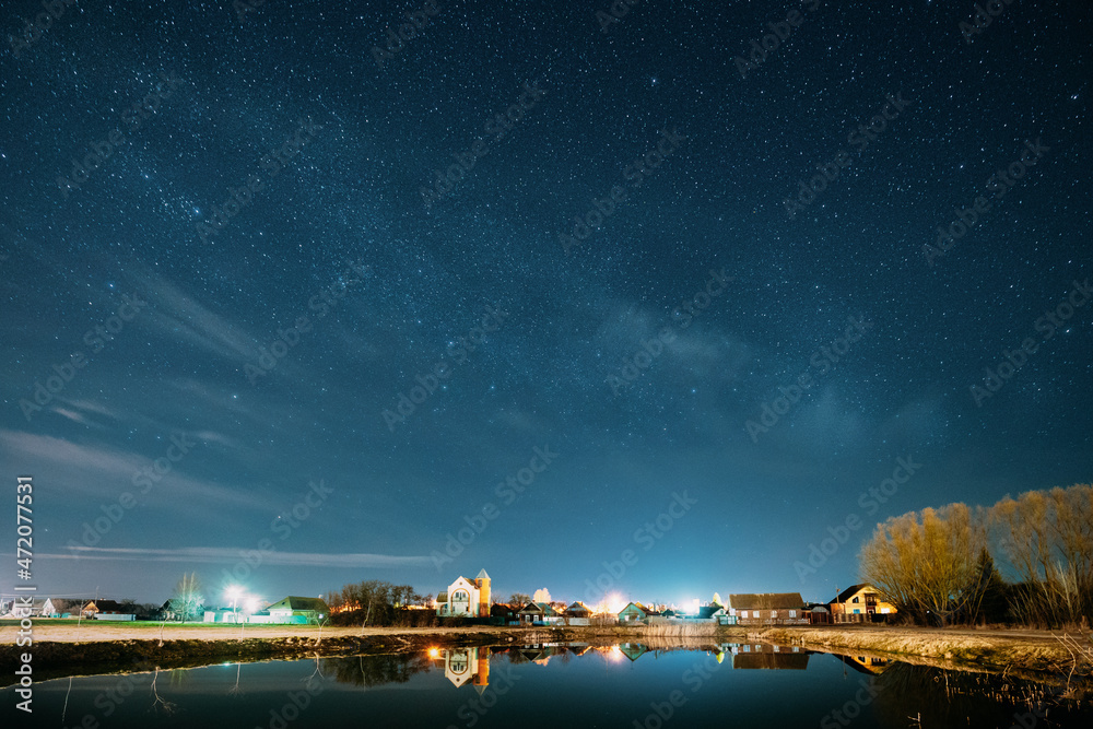 Belarus, Eastern Europe. Night Sky Stars Above Countryside Landscape With Lake Coast And Small Town Or Village. Natural Starry Sky Above Pond And Houses In Early Spring Night Belarus, Eastern Europe. Night Sky Stars Above Countryside Landscape With Lake Coast And Small Town Or Village. Natural Starry Sky Above Pond And Houses In Early Spring Night