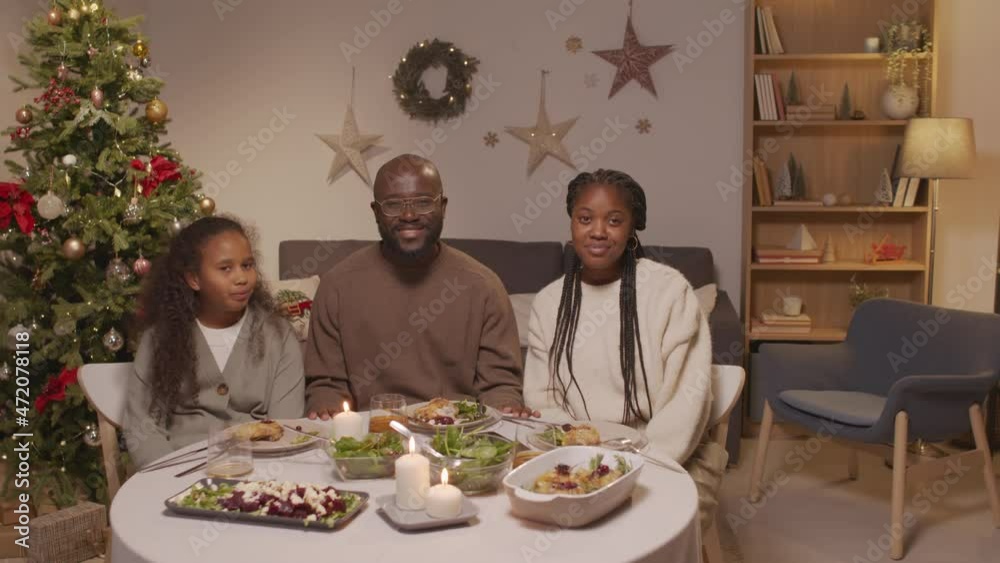 Handheld portrait of happy African-American man, woman and girl sitting at dinner table in living room with Christmas tree and chatting, then posing for camera