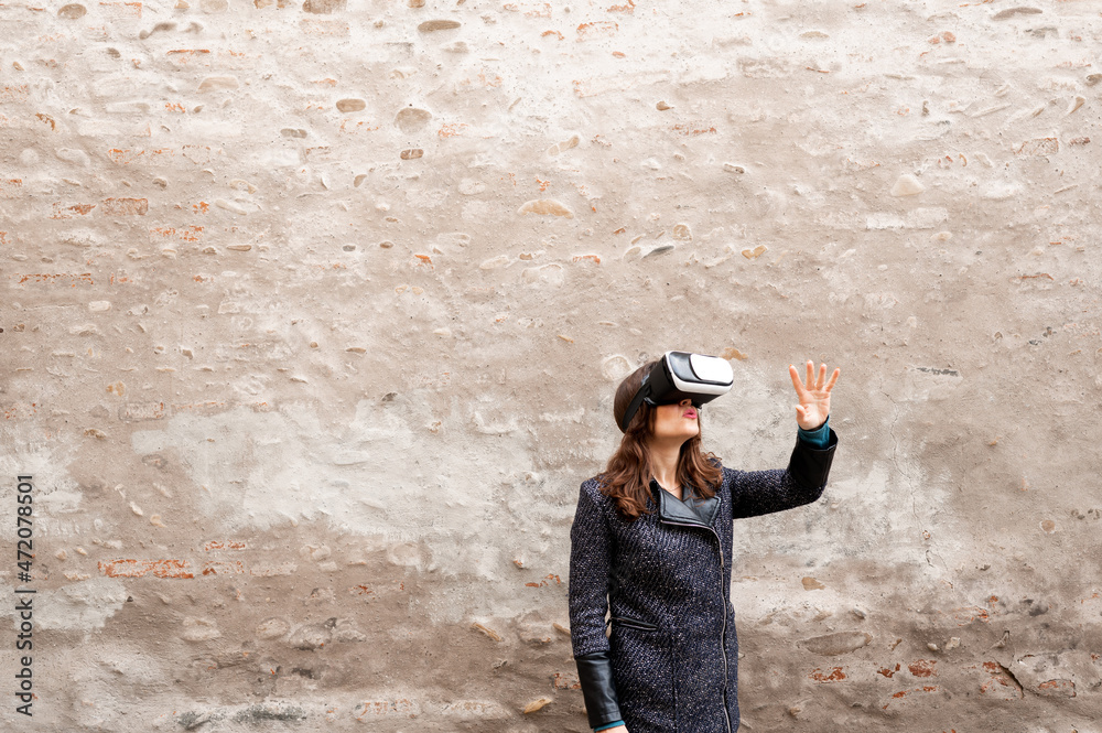 Woman with surprised serious face wearing virtual reality glasses and gesturing with hand. Wide angle lengh with old wall on background and copy space.