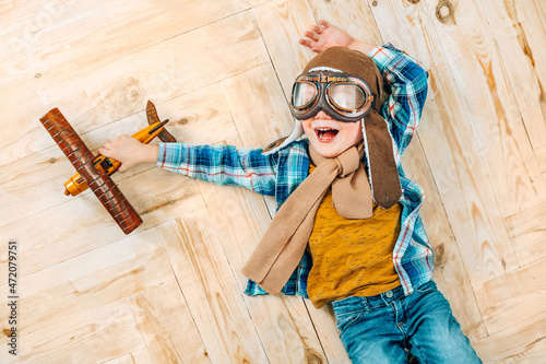 Happy little pilot aviator boy plays with wooden airplane on the floor. Childhood dream and imagination concept.