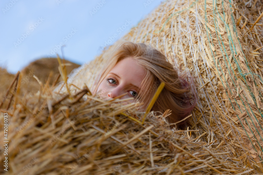 A young beautiful blonde girl with blue eyes and snow-white skin sits on the ground, leaning on a haystack, looks relaxed to the side enjoying the rays of the setting sun in summer. A young beautiful blonde girl with blue eyes and snow-white skin sits on the ground, leaning on a haystack, looks relaxed to the side enjoying the rays of the setting sun in summer.