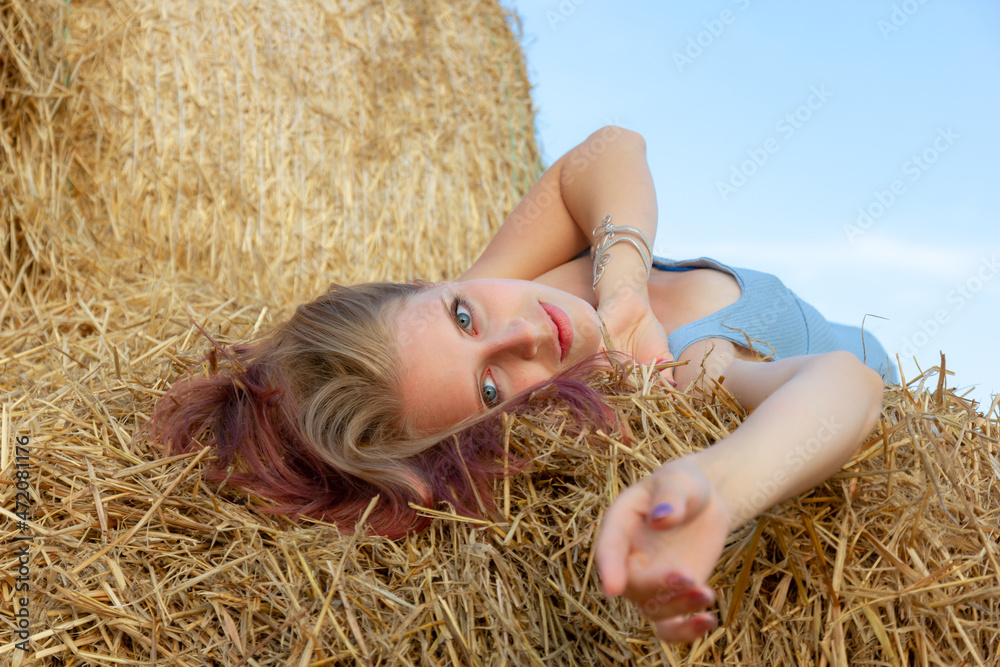 A young beautiful blonde girl with blue eyes and snow-white skin sits on the ground, leaning on a haystack, looks relaxed to the side enjoying the rays of the setting sun in summer. A young beautiful blonde girl with blue eyes and snow-white skin sits on the ground, leaning on a haystack, looks relaxed to the side enjoying the rays of the setting sun in summer.