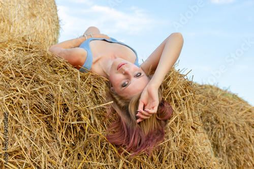 A young beautiful blonde girl with blue eyes and snow-white skin sits on the ground, leaning on a haystack, looks relaxed to the side enjoying the rays of the setting sun in summer.