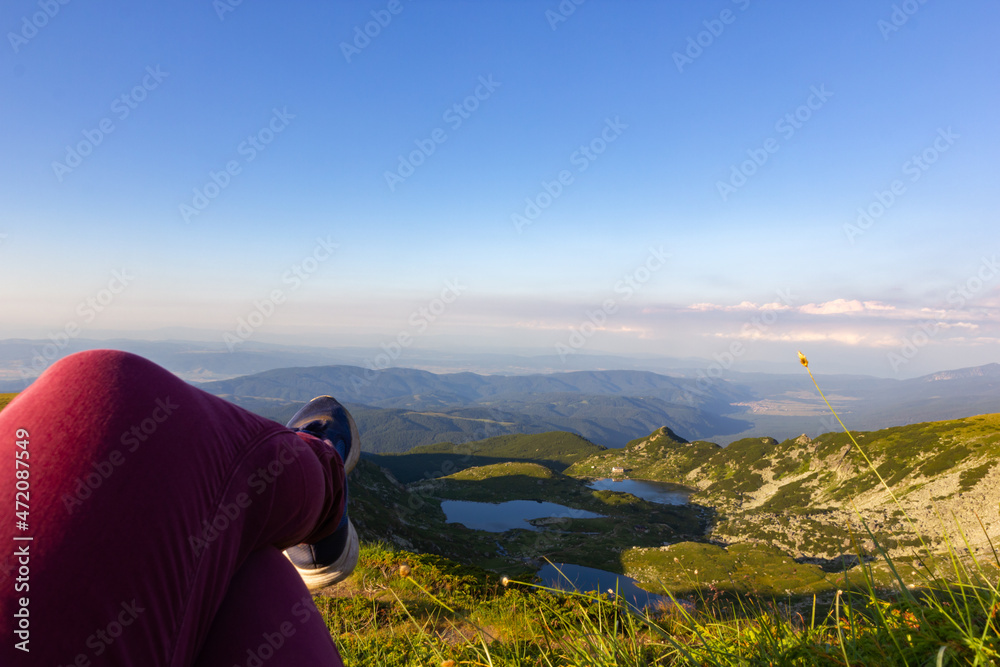 Naklejka premium Against the backdrop of green mountains and mountain lakes and a view of the valley above the clouds, the legs of a human are located in the foreground. Active rest climbing mountains on foot.