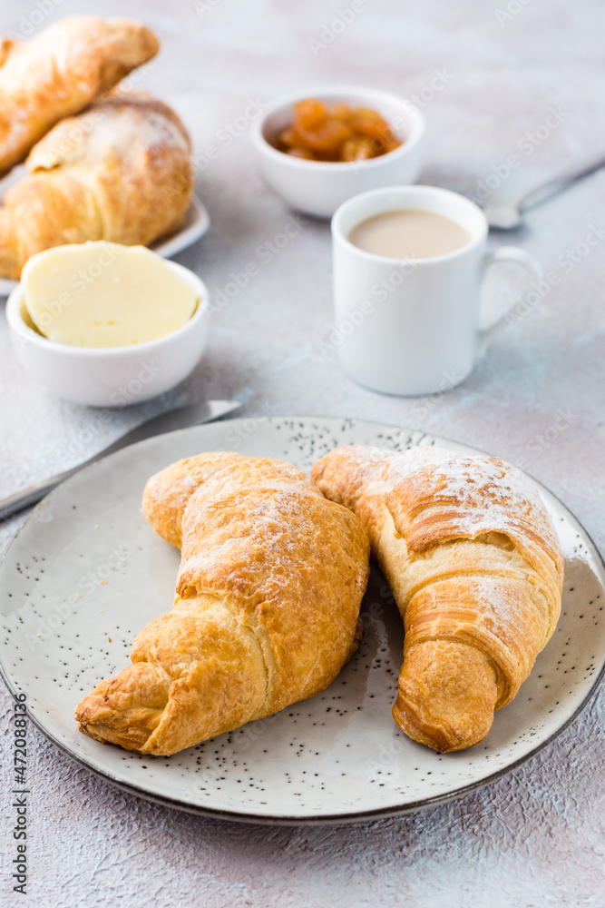 A pair of fresh crispy croissants on a plate, a cup of coffee and bowls of jam and butter on a light textured background. Coffee break or breakfast. Vertical view