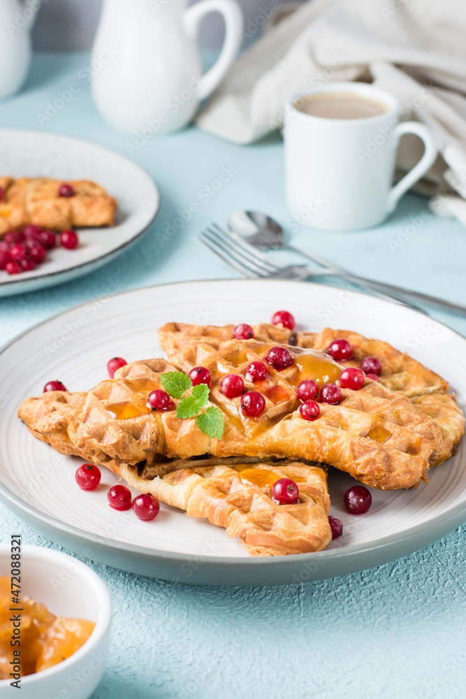 Crispy croissant waffles with red currants, honey and mint on plates and a cup of coffee on a light table. Sweet mouth-watering breakfast - croffles. Close-up. Vertical view