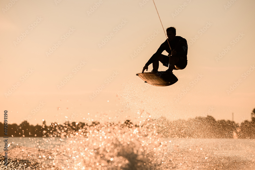 Beautiful silhouette of male rider holding rope and making extreme jump on wakeboard over splashing water.