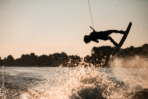 Beautiful silhouette of female rider holding rope and making jump on wakeboard at sunset. Water sports activity.