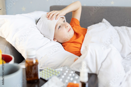 A sick child boy lying in bed covered with a blanket with a fever, near the medicine on the table. Flu season
