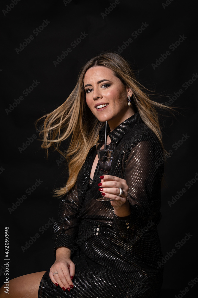 Attractive blonde woman in black sequin and glitter dress with glass in hand looking at camera and smiling on black background in photo studio Attractive blonde woman in black sequin and glitter dress with glass in hand looking at camera and smiling on black background in photo studio
