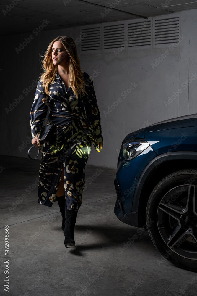 Naklejka premium Elegantly dressed blonde woman walking beside a car at night in a garage