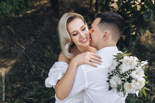 A young, stylish groom gently hugs and kisses a smiling, sweet, curly blonde bride with a bouquet of peonies in her hands in nature. Wedding photography, portrait.