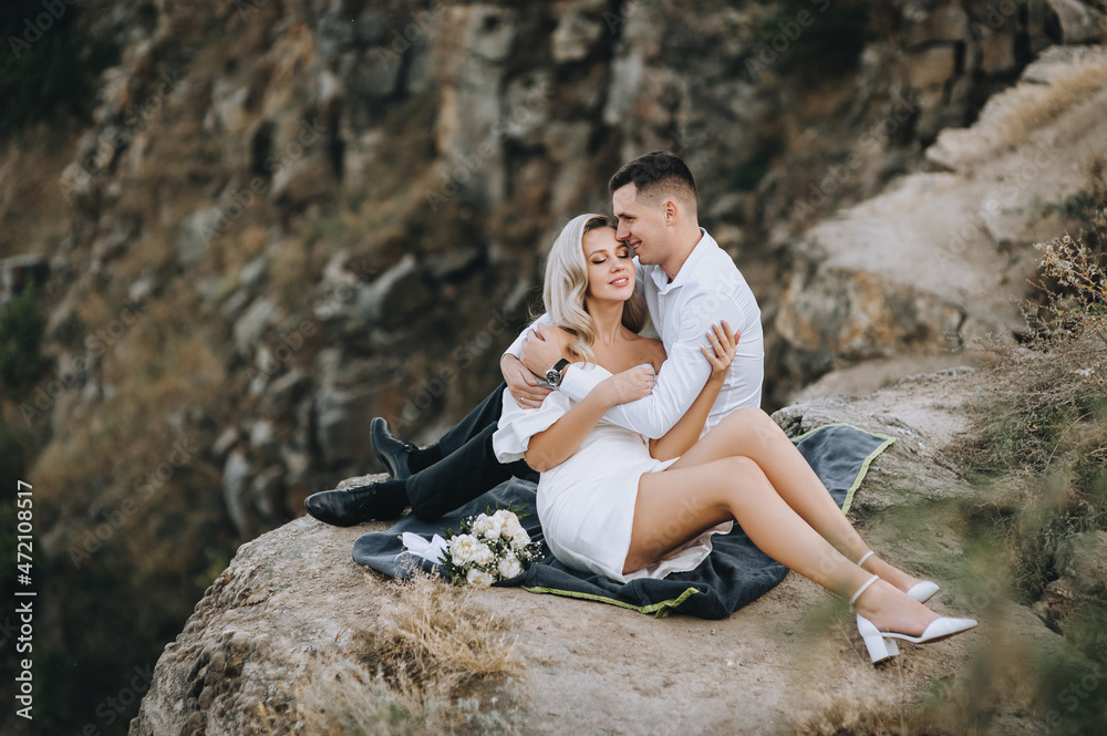 Stylish young groom and a beautiful blonde bride in a white dress are sitting on a rug on a cliff, tenderly embracing, against the backdrop of rocks. Wedding photography.