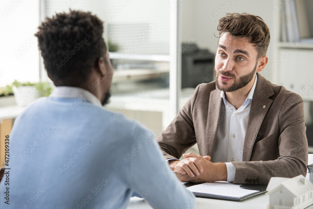 Confident young bearded recruiter sitting at table and asking candidate while holding job interview Confident young bearded recruiter sitting at table and asking candidate while holding job interview