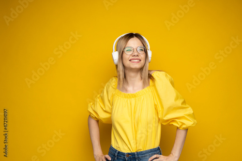 Blonde woman in a glasses and yellow shirt listening music with a headphones. Teenager girl dancing against yellow background. © Volodymyr