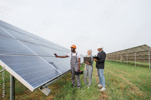 African american technician measuring temperature of solar panels with thermal imager. Muslim woman in hijab typing on laptop, indian man writing on clipboard. Inspection of station.