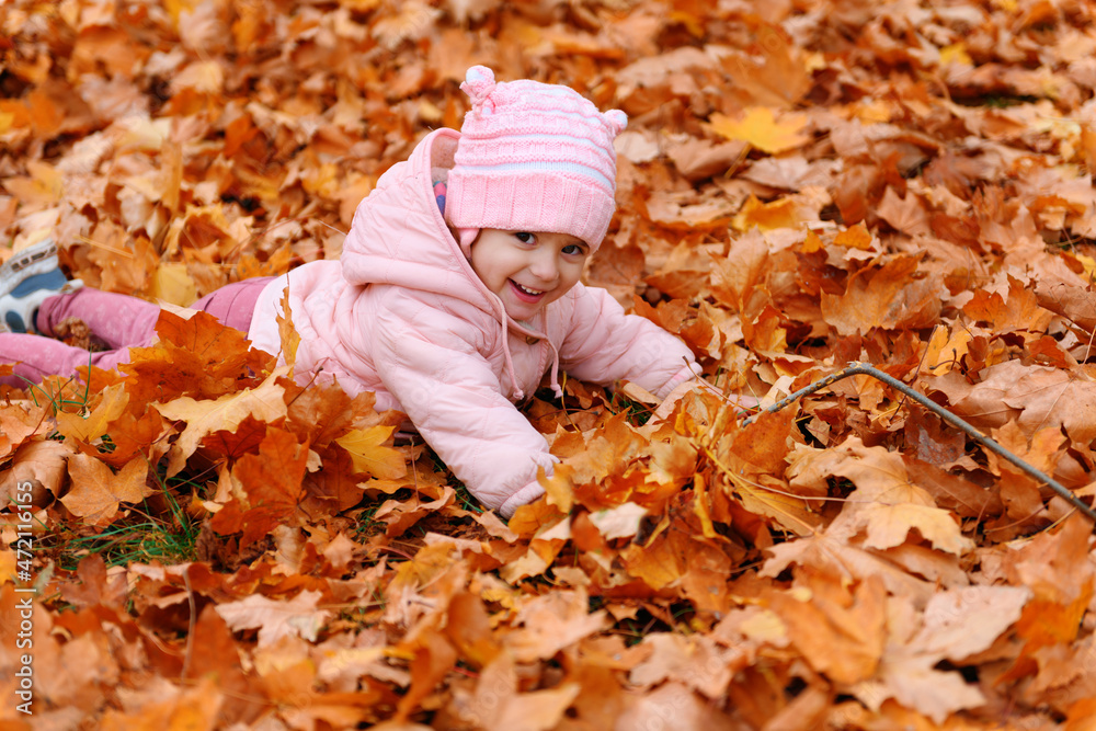 Child girl sitting on fallen leaves in autumn city park. Beautiful nature, trees with yellow leaves.