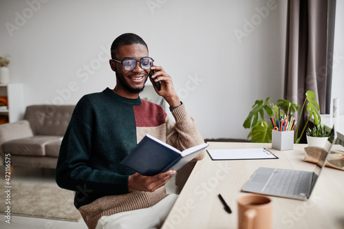 Portrait of young African-American man wearing glasses while working from home and speaking by smartphone, copy space