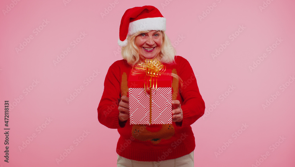 Senior grandmother old woman wears red New Year sweater presenting Christmas gift box stretches out hands isolated on pink studio wall background. Happy Christmas celebration holiday shopping sale eve