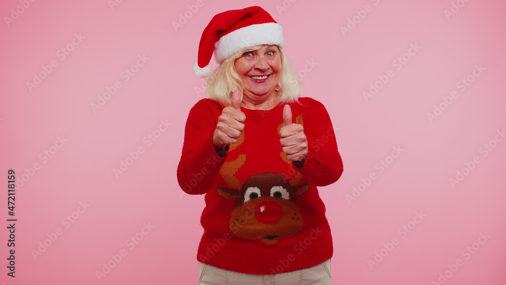 Senior grandmother in New Year sweater with deers raises thumbs up agrees with something or positive reply recommends advertisement likes good on pink background. Happy Christmas celebration holidays