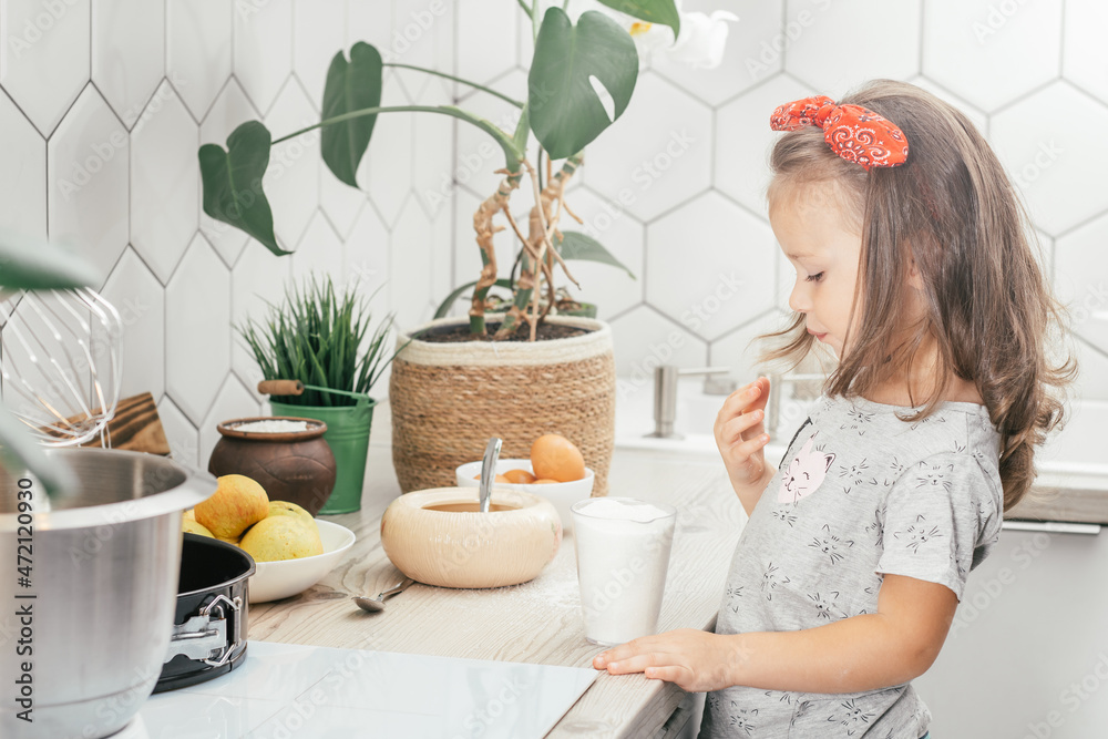 Little dark-haired girl 3 years old in red headband bakes apple pie in kitchen. Child puts sugar in measuring cup