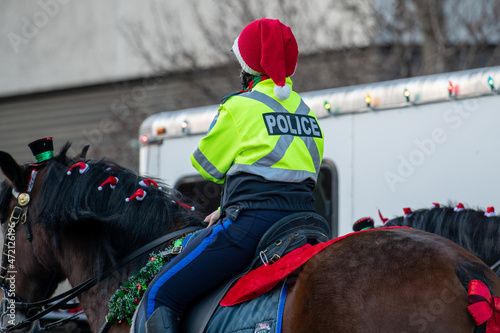 A female police officer wearing a fluorescent yellow coat with the word police on the back and a red Santa Claus hat rides a large chestnut brown horse decorated with Christmas garland, bows, and hat.