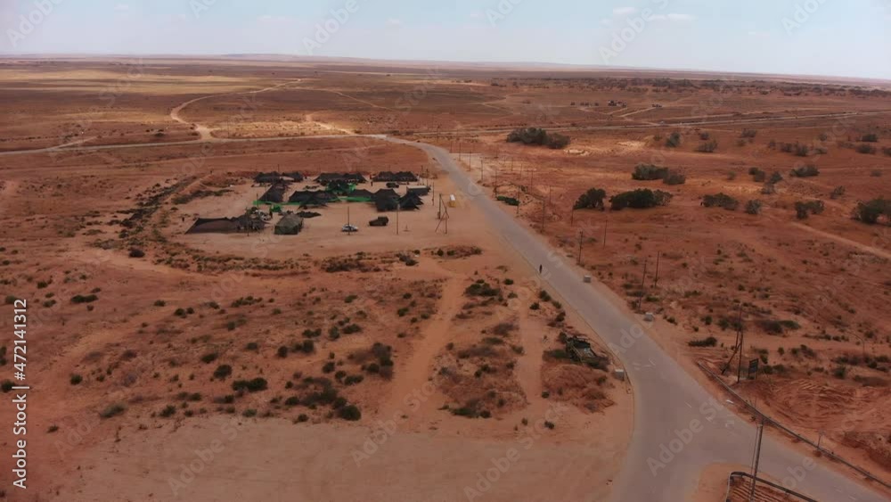 aerial static shot of army camp of tents in the middle of the endless desert next to an empty road with single soldier walks in it.