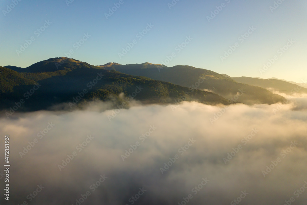 Aerial view of vibrant sunrise over white dense fog with distant dark silhouettes of mountain hills on horizon