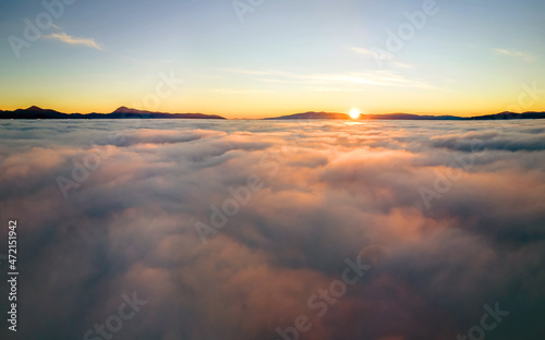 Aerial view of vibrant yellow sunrise over white dense clouds and distant mountains on horizon