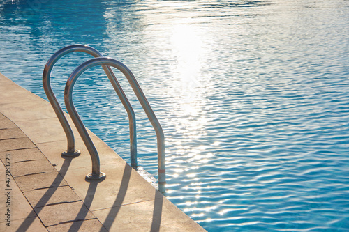 Close up of swimming pool stainless steel handrail descending into tortoise clear pool water. Accessibility of recreational activities concept © bilanol