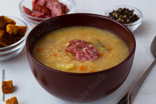 The brown bowl of vegetable soup with meat and crackers. Split pea soup with ham, carrots and potatoes with spoon on a rustic white background. Classic Dutch pea soup erwtensoep, snert with bacon.